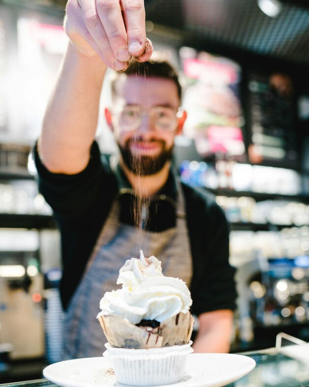 Cafè-Mitarbeiter streut Kakaopulver auf Cupcake | unique by ATLANTIC Hotels