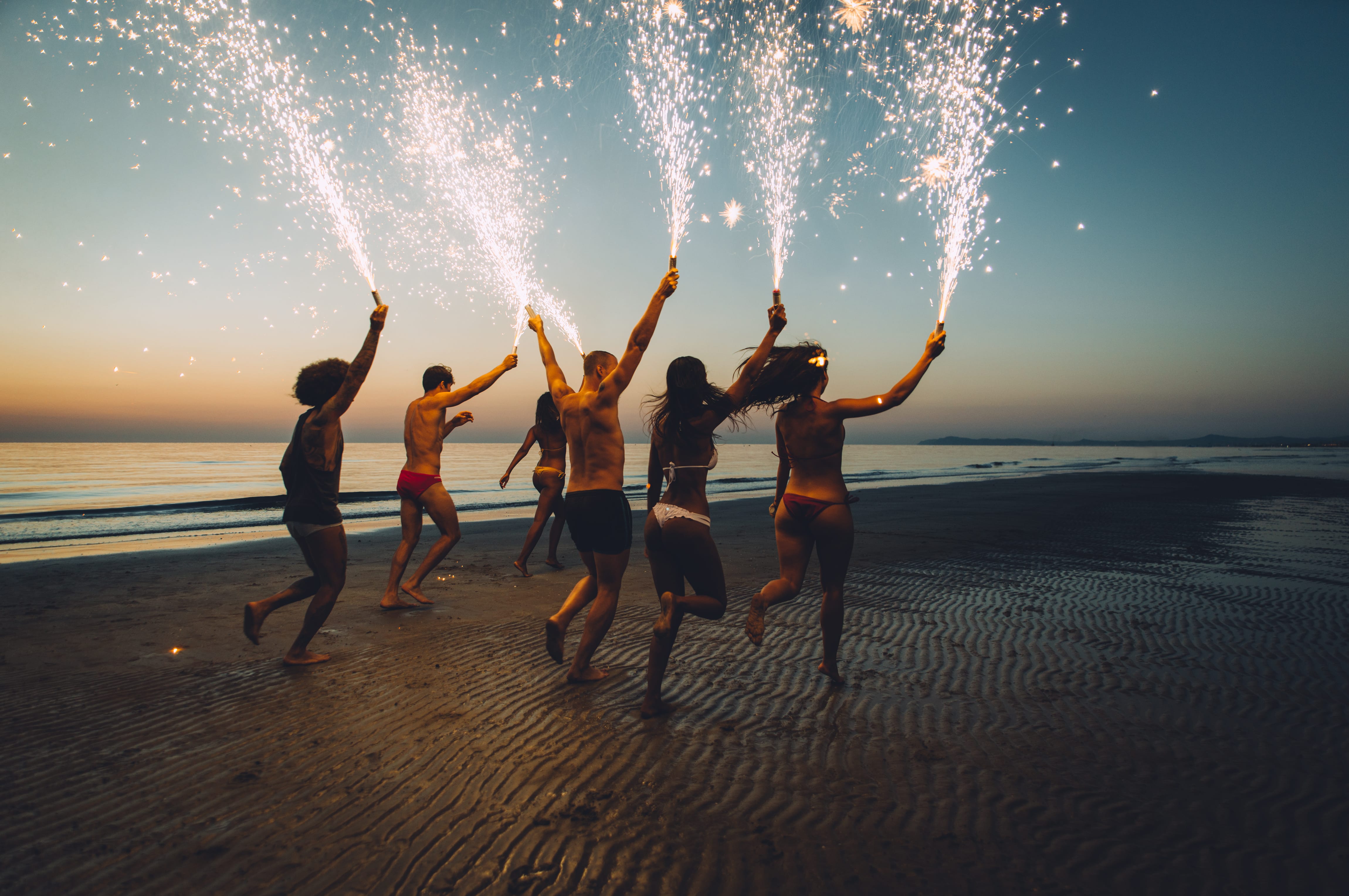 Gruppe in Badesachen und mit Feuerwerksfontänen in der Hand laufen am Strand | unique friends