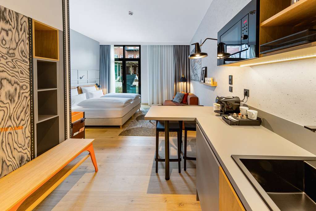 Modernes Hotelzimmer mit Doppelbett, Küchenzeile, Sitzbereich und großen Fenstern mit Blick auf eine Terrasse.