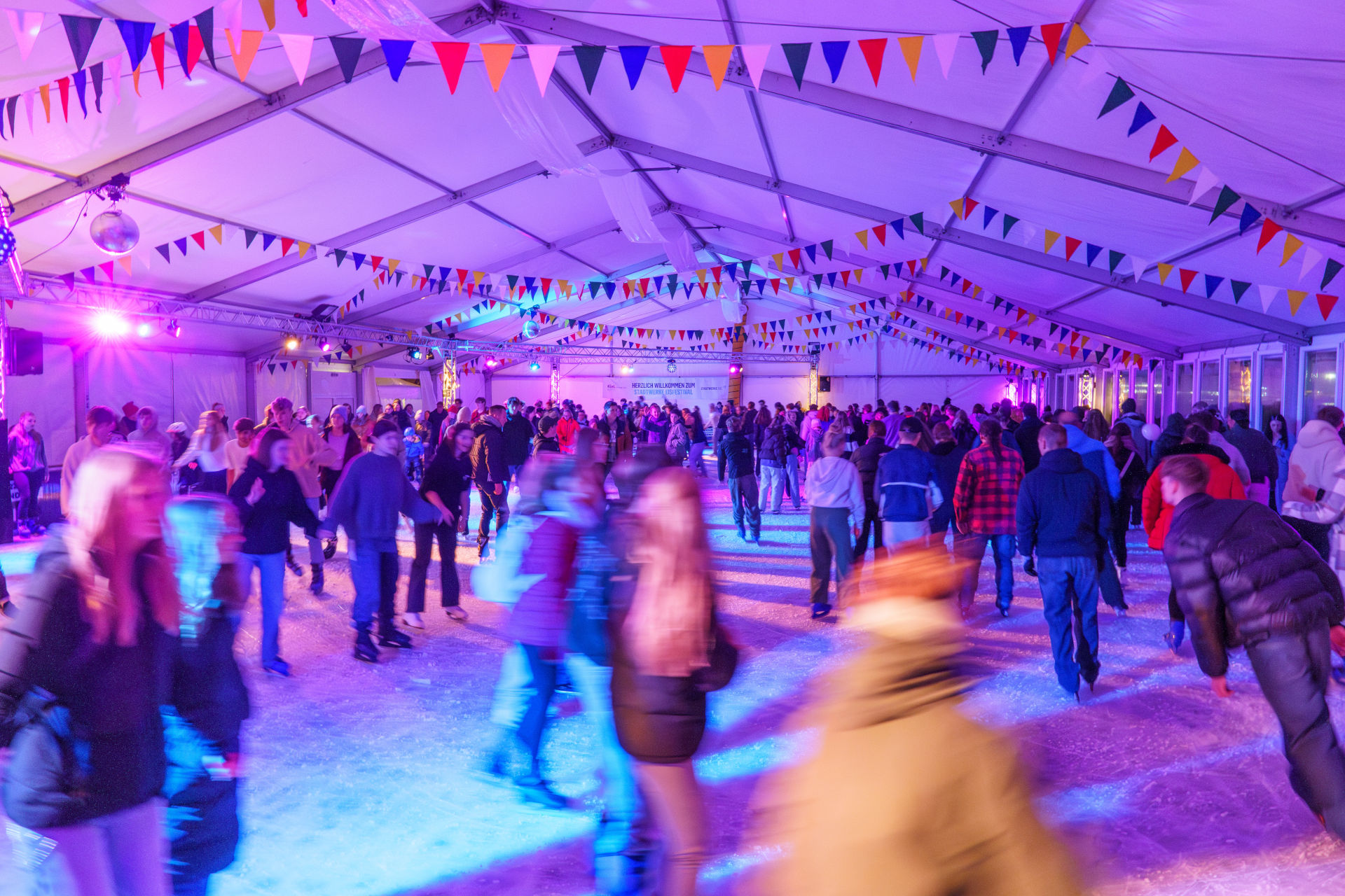 Eislaufbahn in Zelt mit bunten Lichtern und Wimpeln, viele Menschen beim Schlittschuhlaufen.