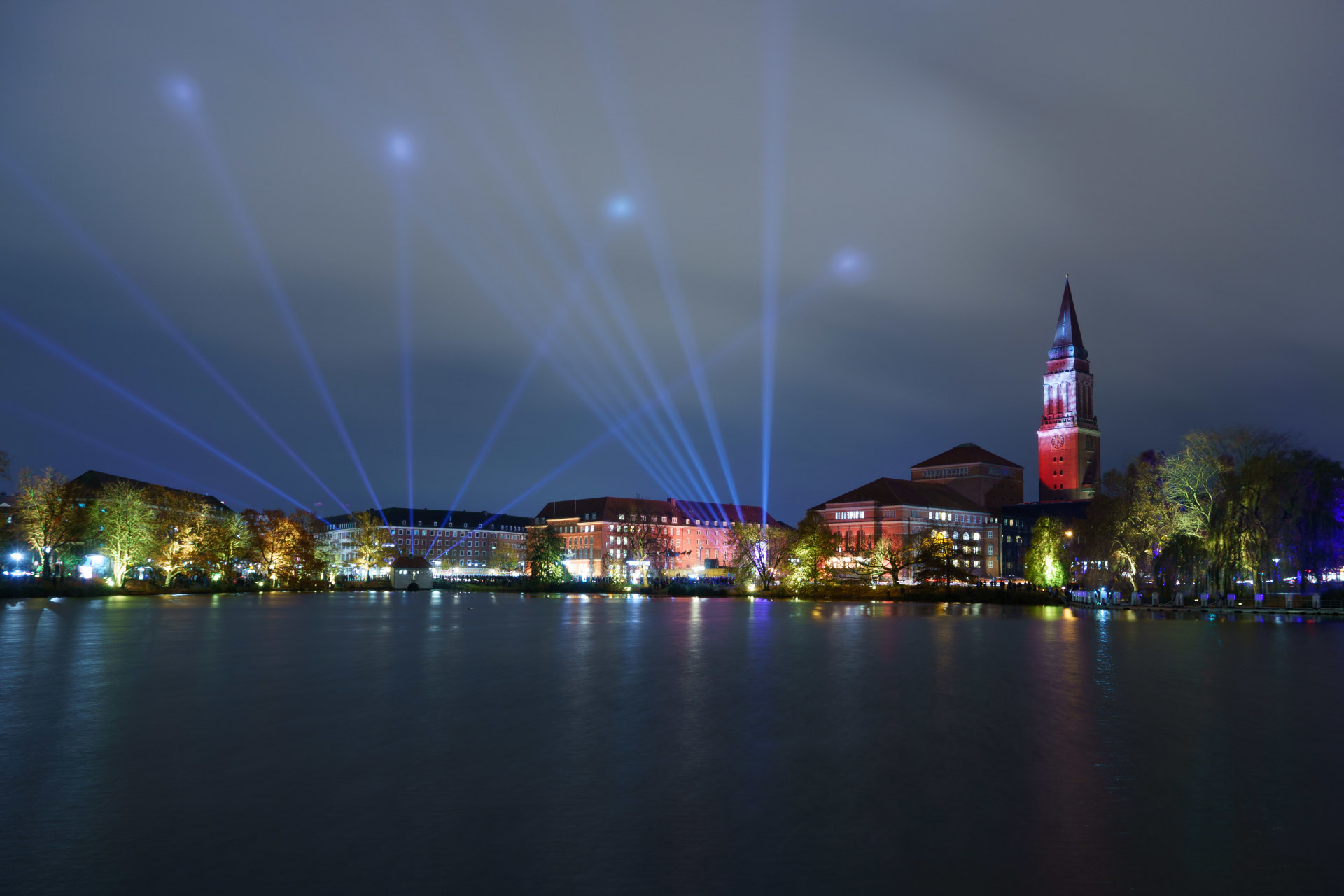 Nachtaufnahme der Kieler Förde mit beleuchteten Gebäuden und Laserstrahlen am Himmel.