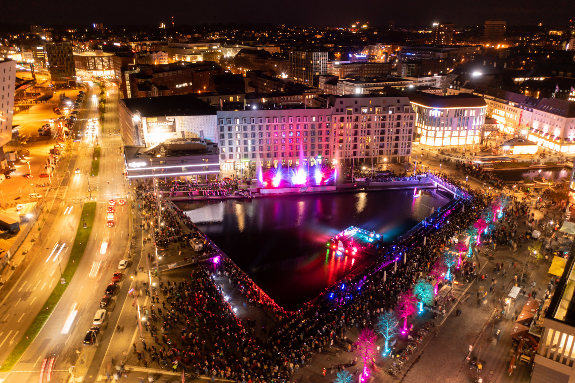 Lichtermeer in Kiel | unique by ATLANTIC Hotels Kiel Nachtaufnahme von beleuchtetem Hotel am Hafen, mit bunten Lichtern und Menschenmengen bei einem Event.