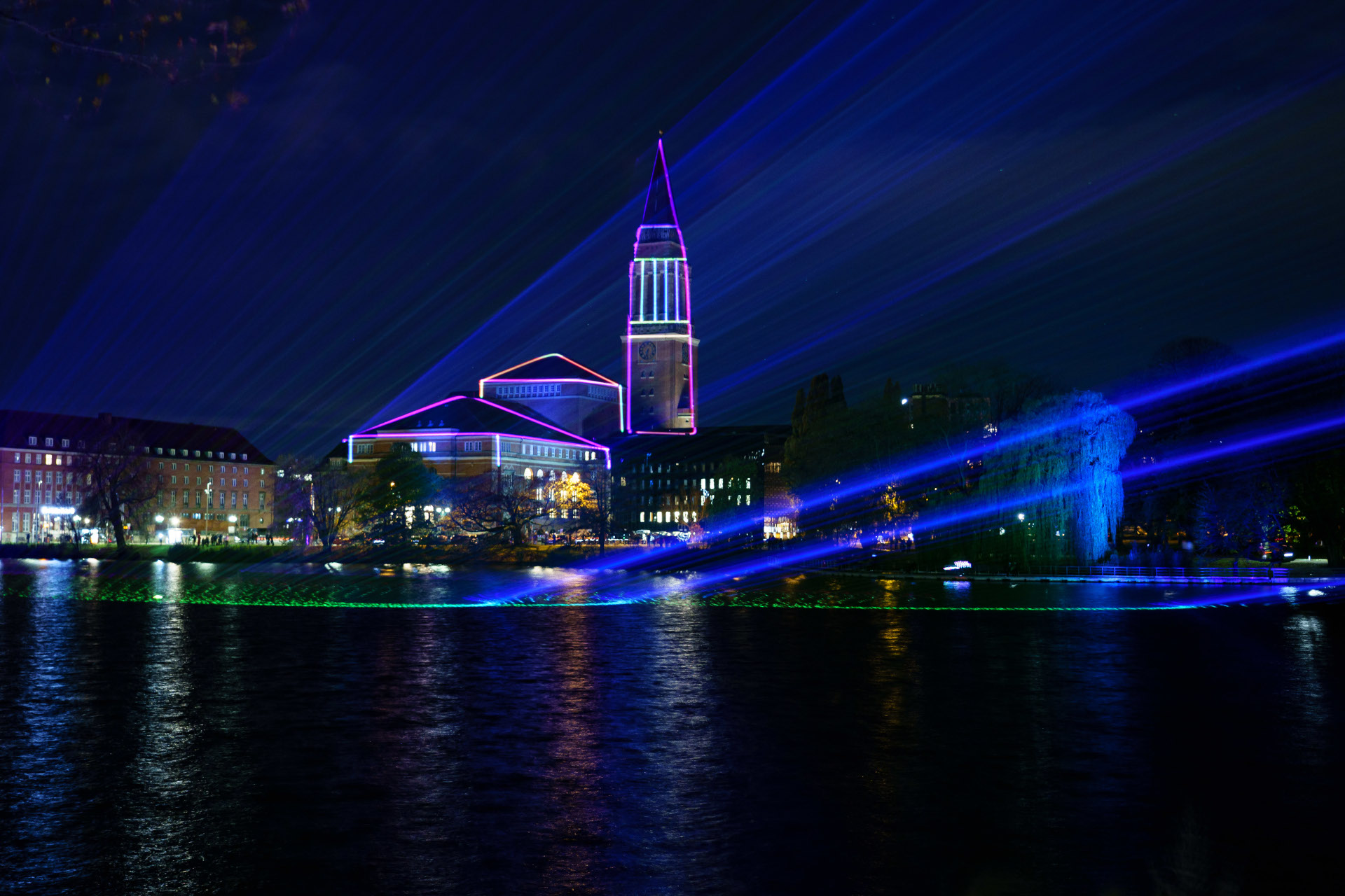 Lichtermeer in Kiel | unique by ATLANTIC Hotels Kiel Nachtaufnahme von Kiels Rathaus mit farbiger Beleuchtung und Lichtstrahlen über Wasser.