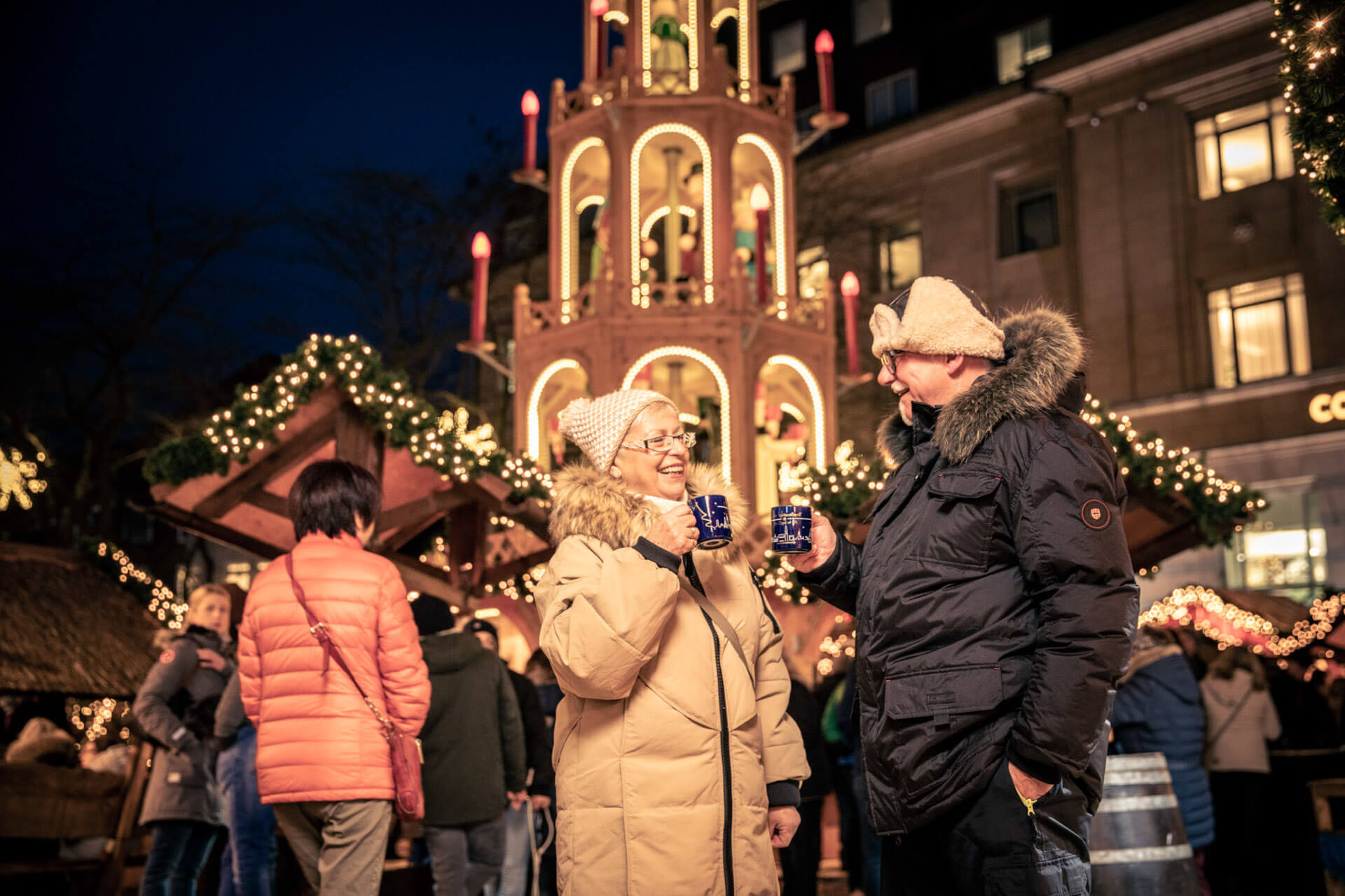 Mann und Frau prosten sich mit einer Weihnachtstasse zu Weihnachtsmärkte in Kiel | Buden mit heißen Getränken